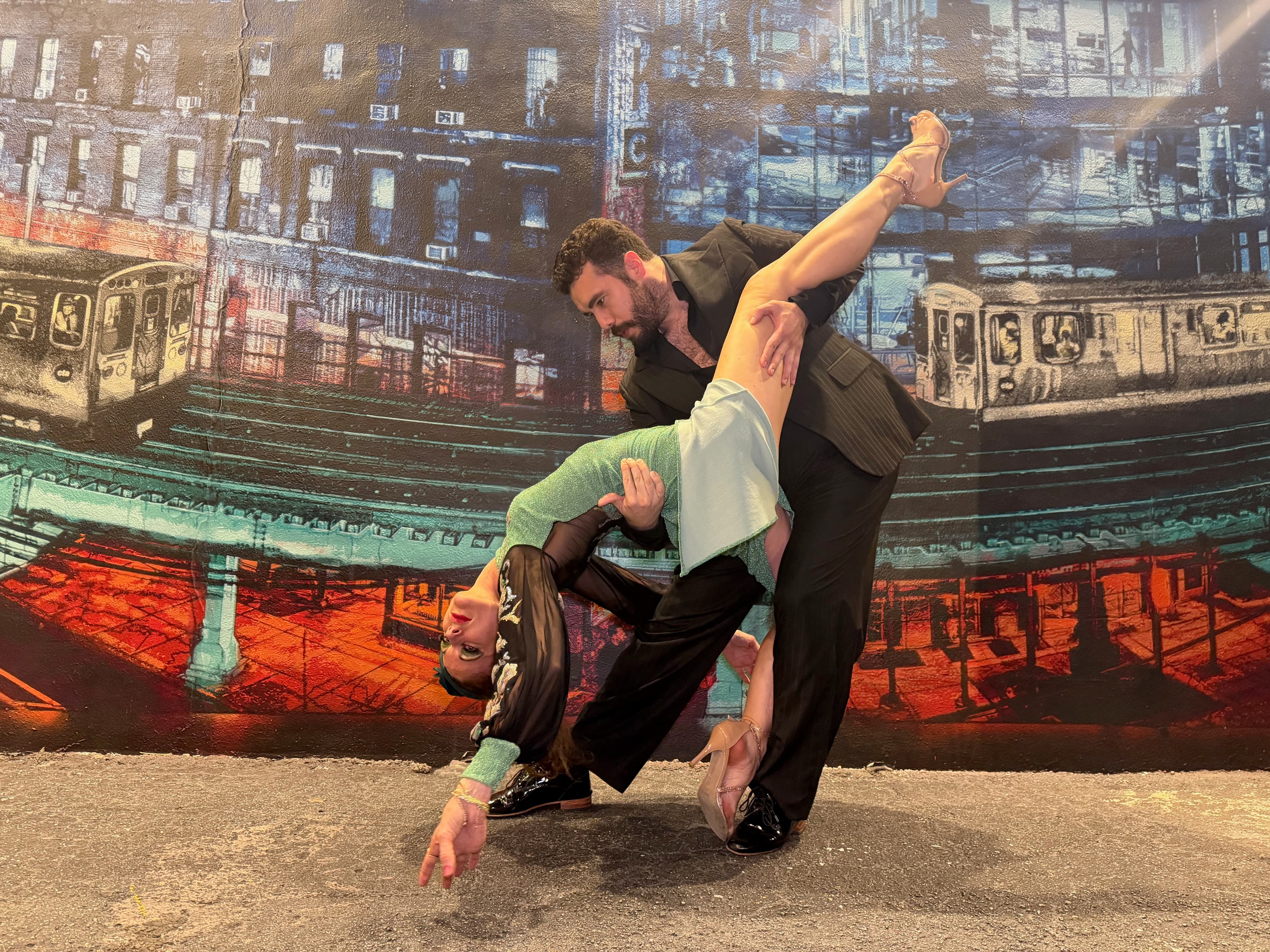 Man in a suit dips a woman in a green dress against a subway mural.